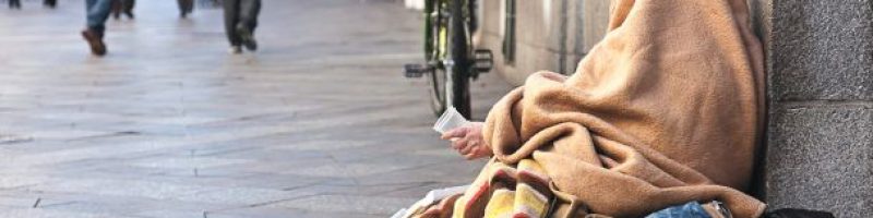 person covered by a blanket while sitting on the sidewalk leaning back against a building